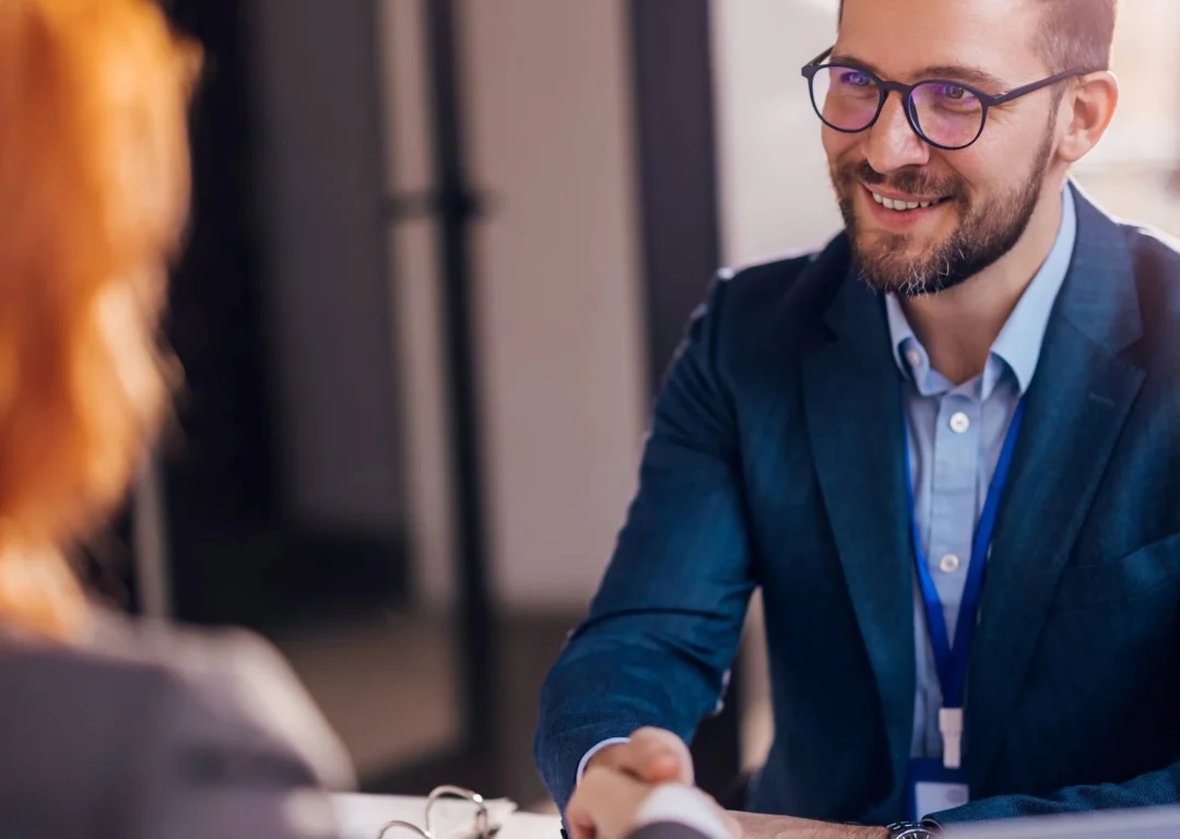 A man wearing a suit and glasses shakes his client's hand.