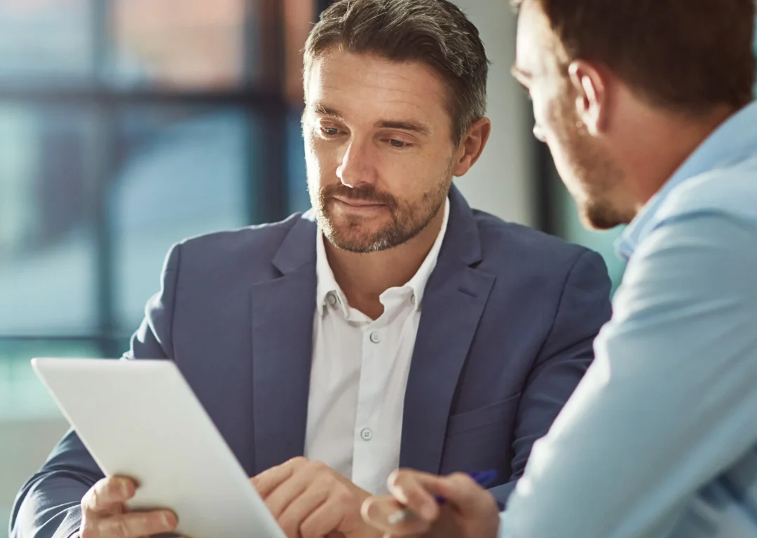 Two men sit at a desk and negotiate using a tablet.
