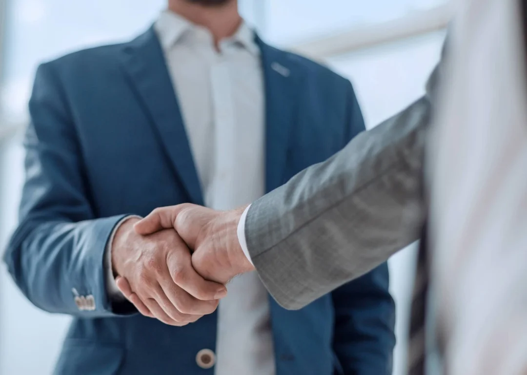 Two men wearing blue and grey suits shake hands.